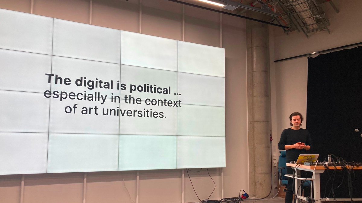 Robert Schnüll on the right, standing at a desk looking at his computer screen in front of him. The majority of the picture of the left is taken by the whiteboard showing the presentation slides, with the current one saying: “The digital is political … (linebreak) especially in the context of art universitites.”